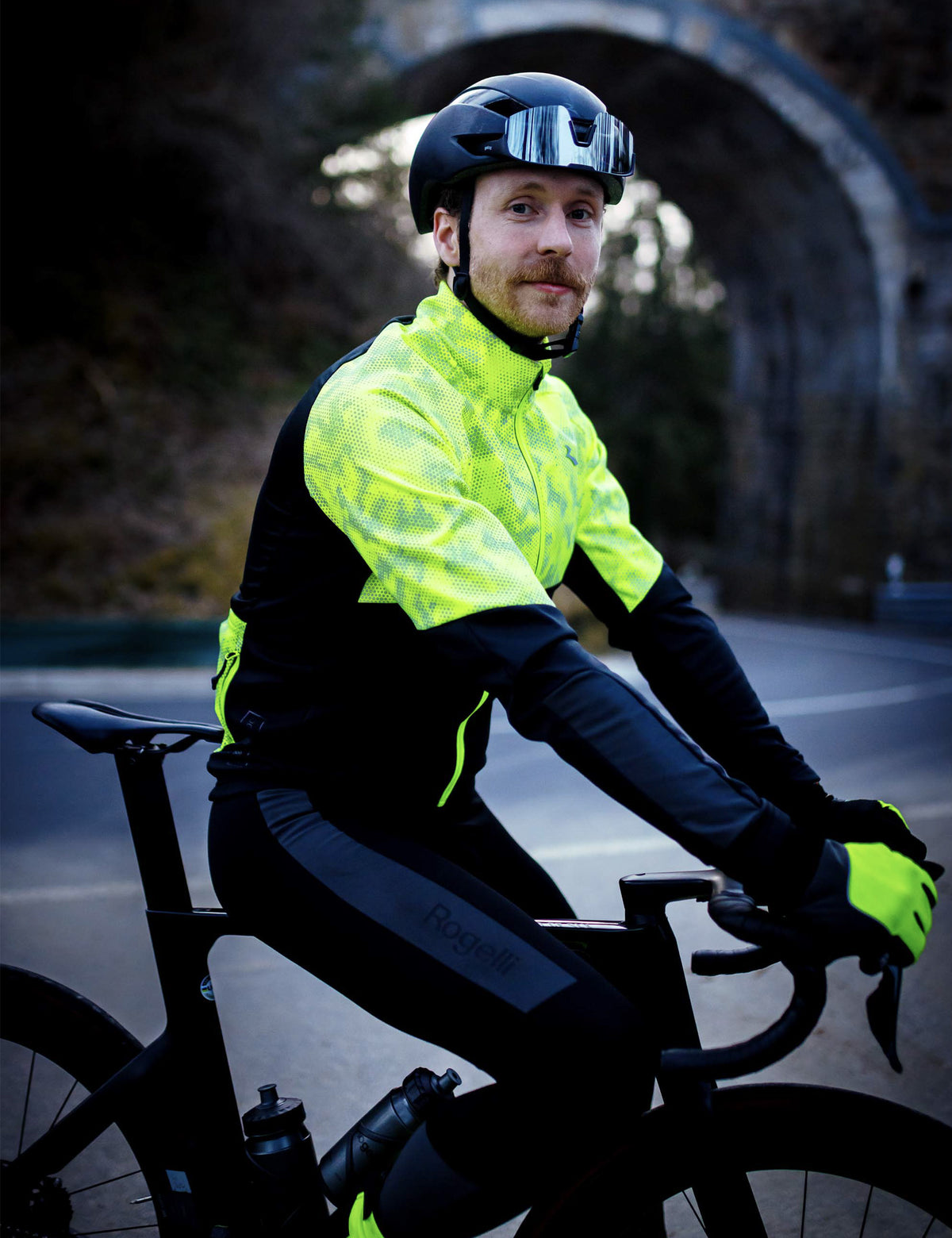 Man op racefiets in Rogelli Essential Hi-Vis winter fietsjack voor heren in fluorgeel, zichtbaar tijdens een rit in donkere omstandigheden.