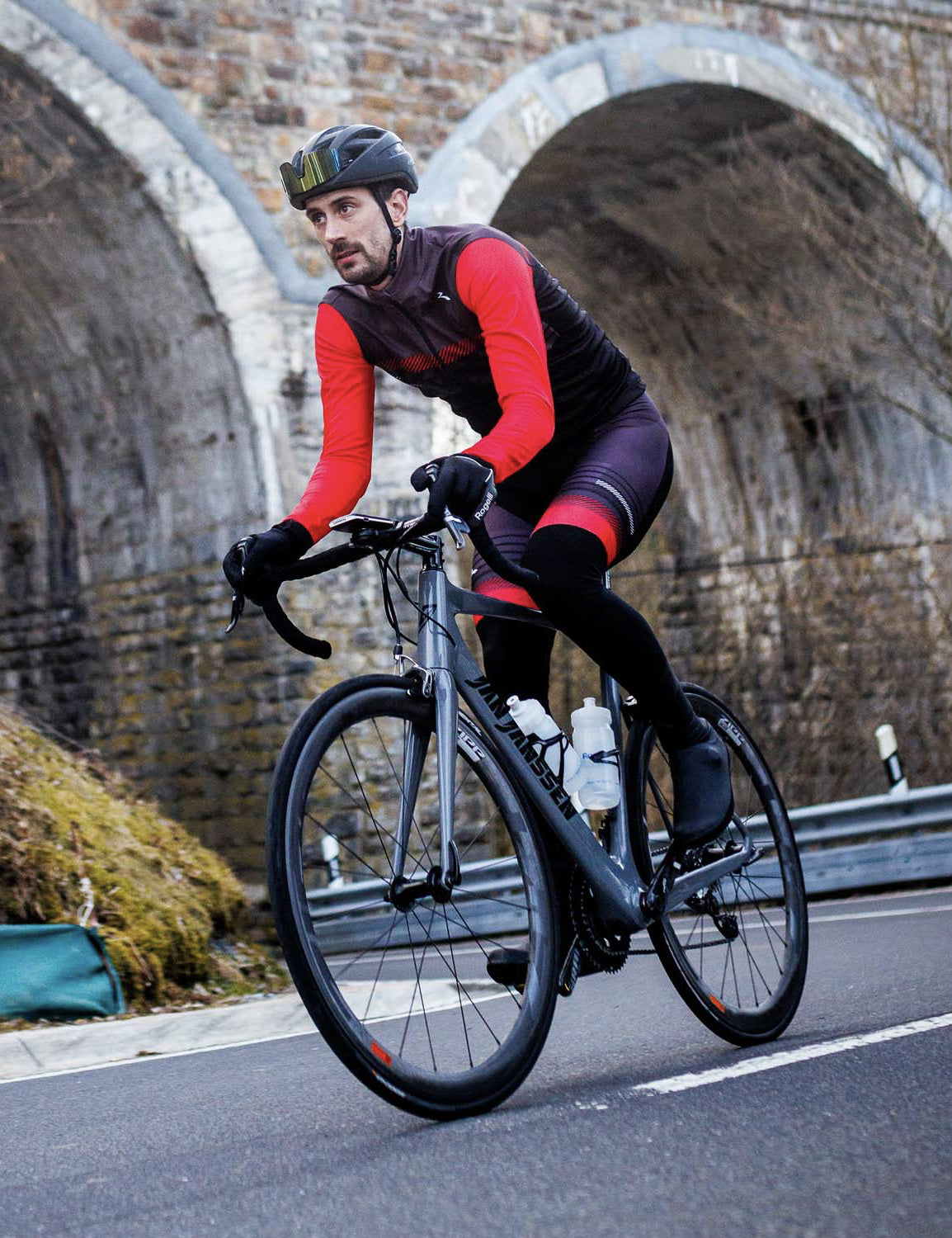 Man in Rogelli Stryde winter fietsjack heren in zwart, grijs en rood op de racefiets in een bochtige buitenomgeving.