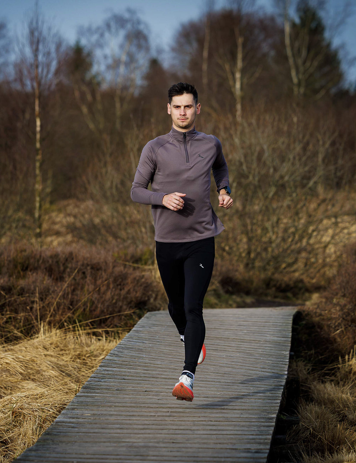 Man in Rogelli hardloopkleding loopt over een houten vlonderpad door een natuurlijke, bosrijke omgeving.