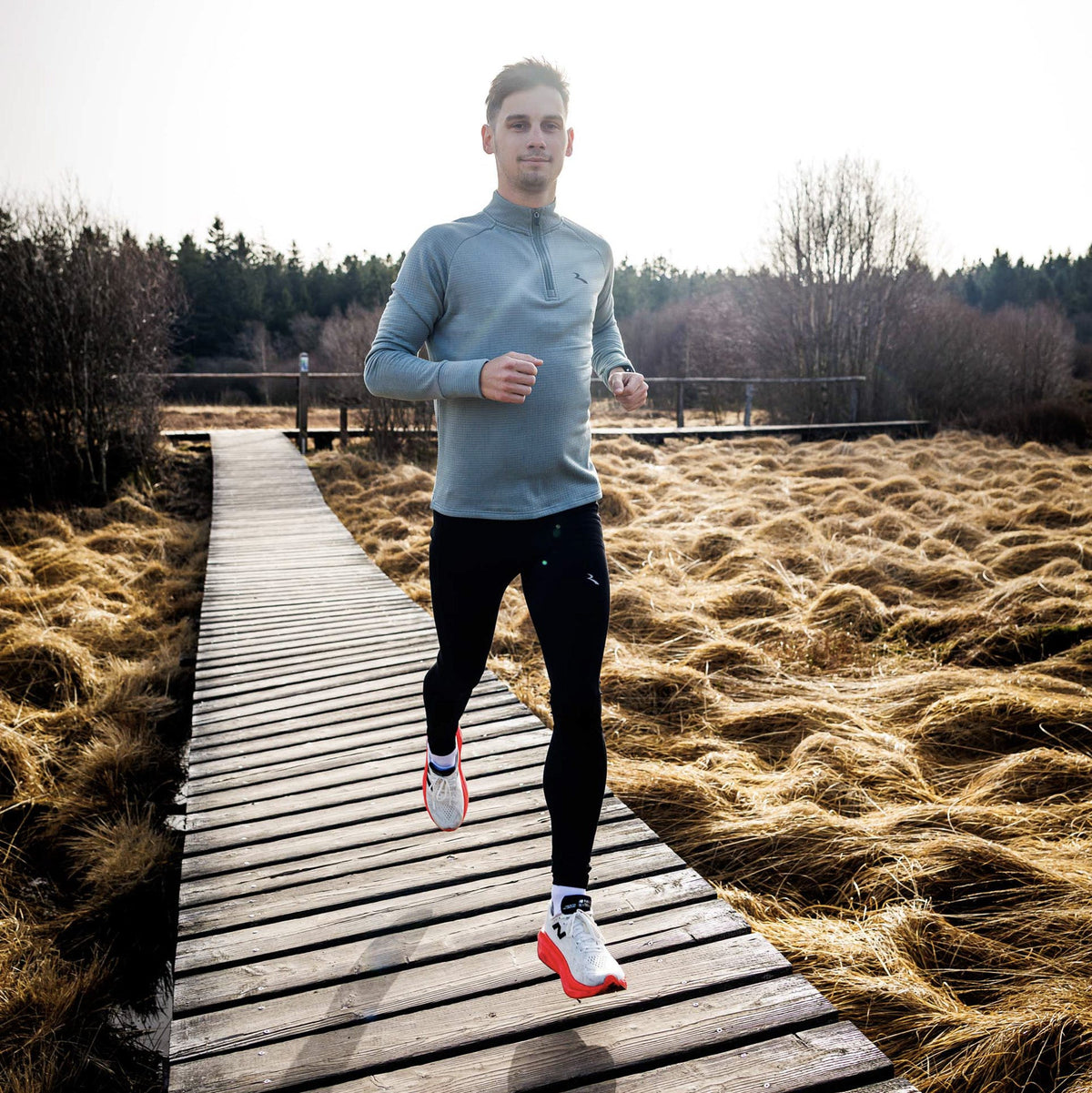 Hardloper in grijsblauwe hardlooptop met halve rits en zwarte lange tight rent over een houten pad door een winters landschap.