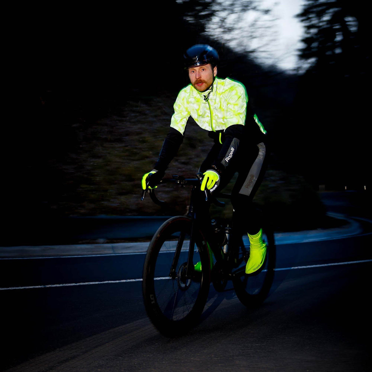 Wielrenner in fluorgeel en zwart winterjack met reflecterende details, fietsend in het donker op een weg door het bos.