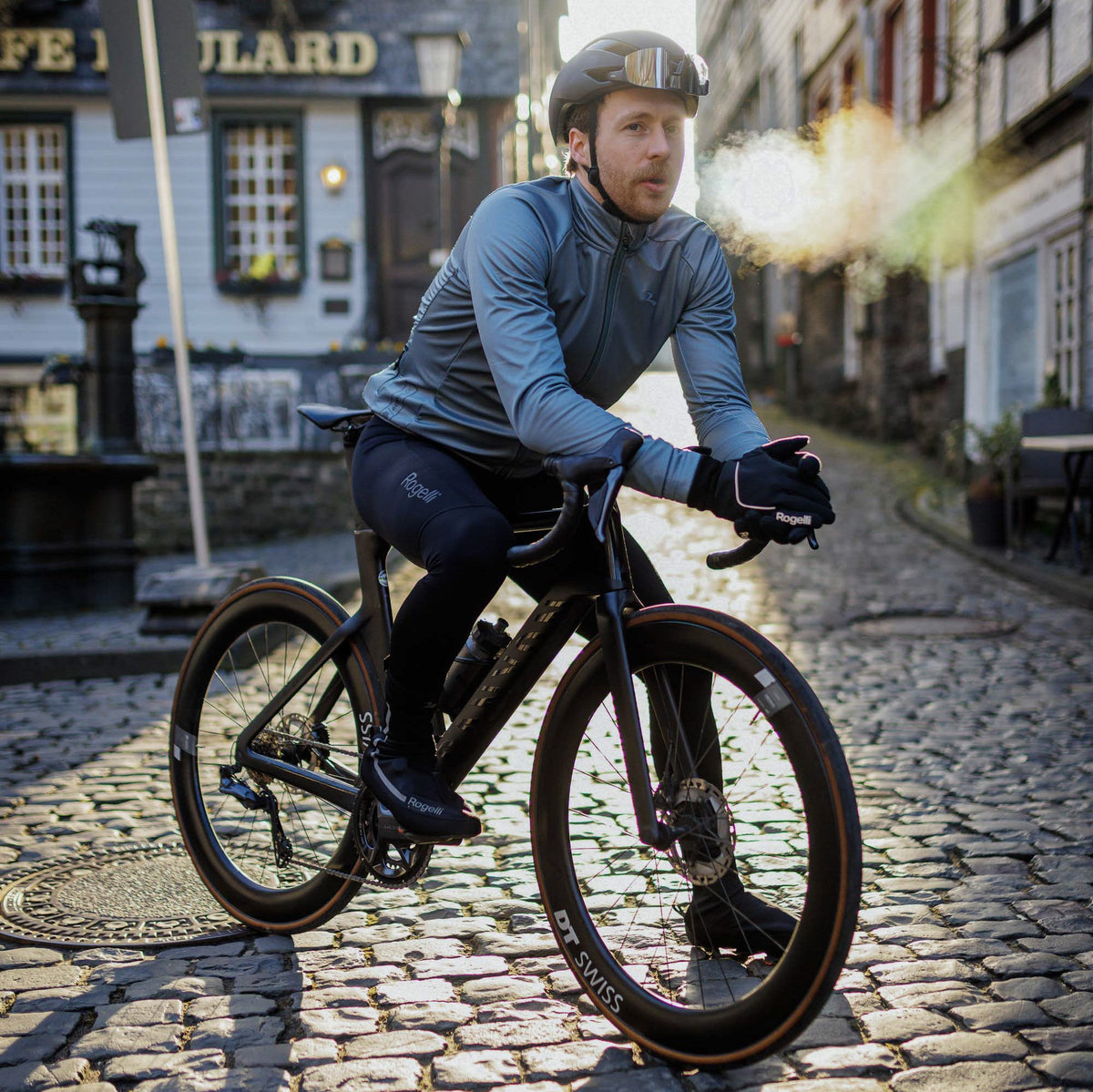 Wielrenner in grijsblauw winterjack, lange fietsbroek en handschoenen van Rogelli op kasseien, met zichtbare adem in de kou.