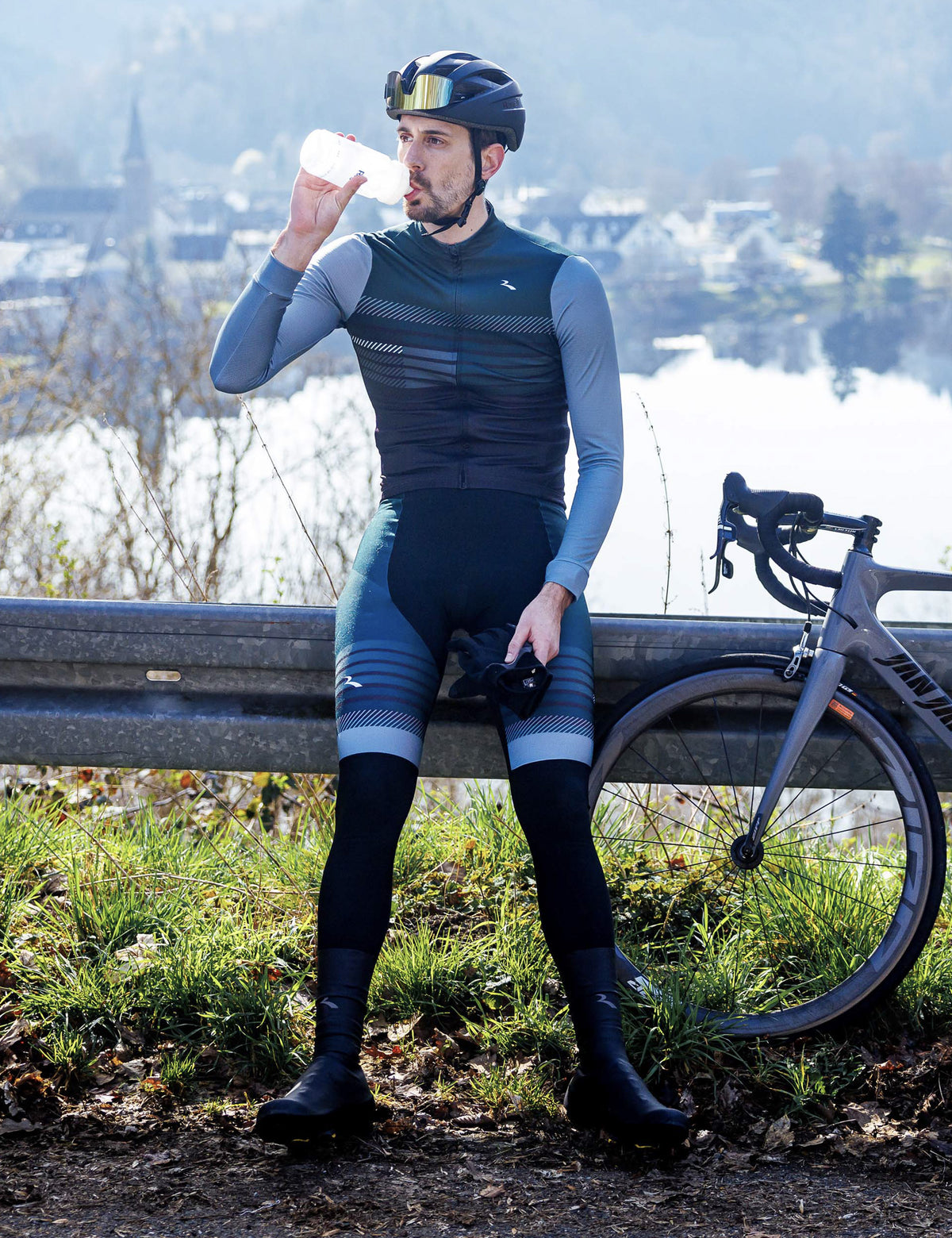 Man in zwarte Rogelli Stryde bibtight met blauw-grijze accenten staat naast zijn racefiets en drinkt uit een bidon tijdens een pauze.