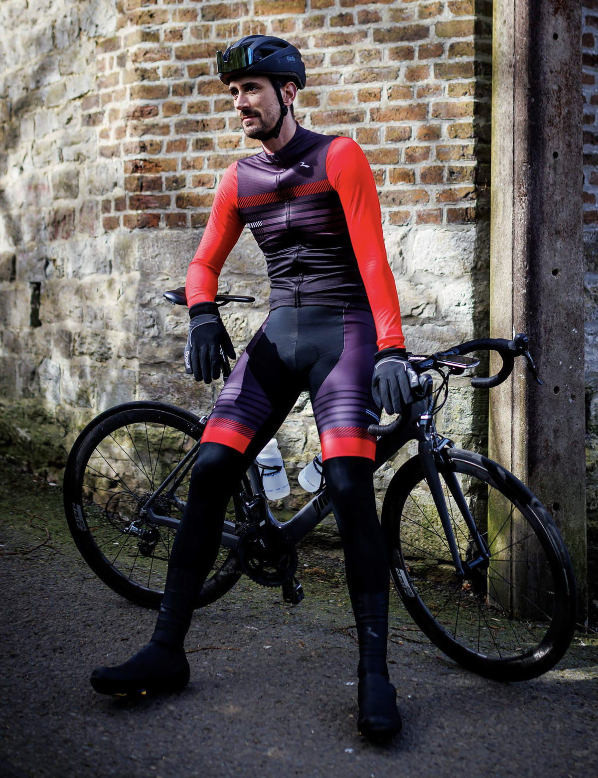 Man in zwarte Rogelli Stryde bibtight met rood-grijze accenten leunt tegen een racefiets bij een bakstenen muur.