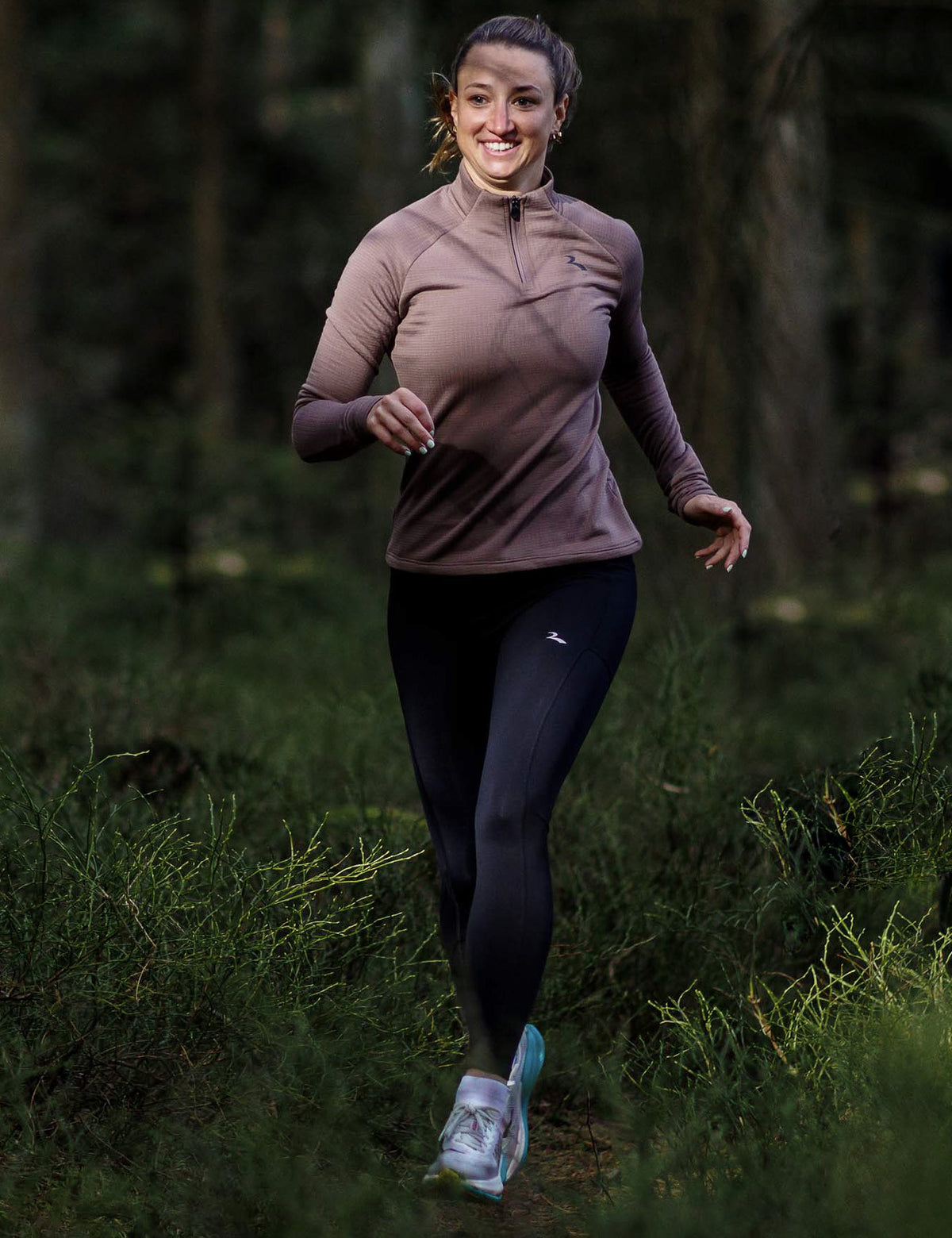Vrouw in Rogelli hardloopshirt en hardlooplegging loopt over een smal bospad door een groene, bosrijke omgeving.