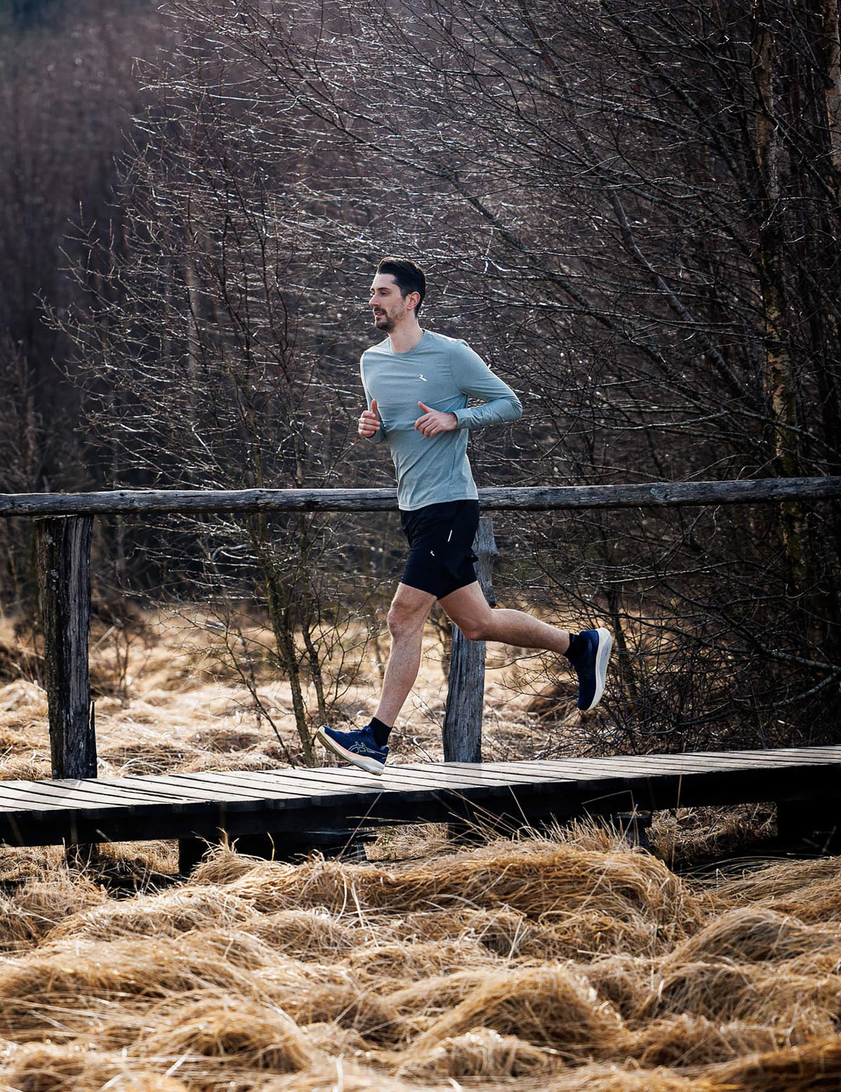 Man in slate Rogelli Essential hardloopshirt heren met lange mouwen tijdens het hardlopen in een bosrijke omgeving.