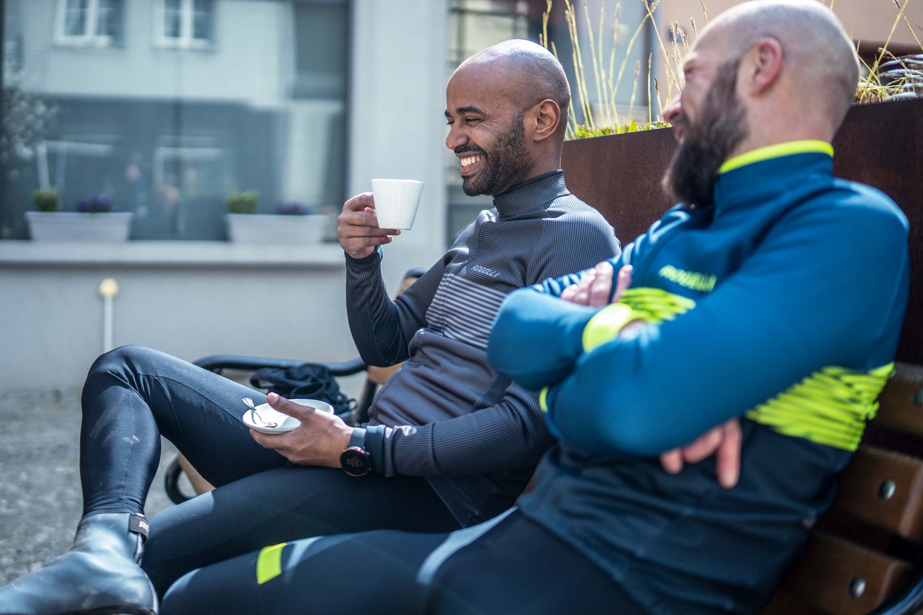 Twee mannelijke wielrenners ontspannen en genieten van een gezellig moment met koffie op een bankje in Luxemburg, terwijl ze samen lachen en plezier maken tijdens hun rustmoment.