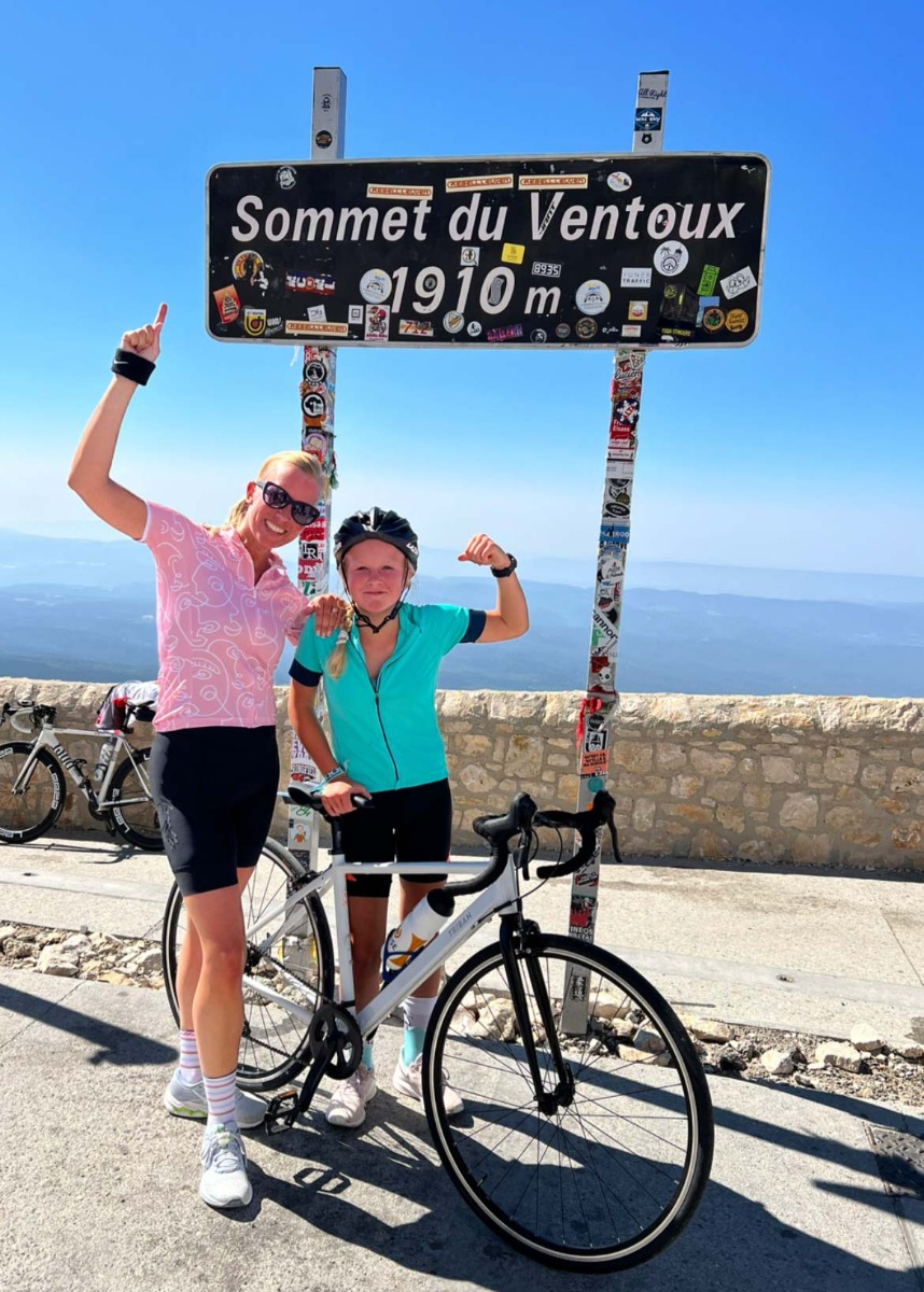 Met een glimlach van voldoening poseren twee wielrenners bij het hoogste punt van Mont Ventoux, een moment van triomf na een uitdagende beklimming.