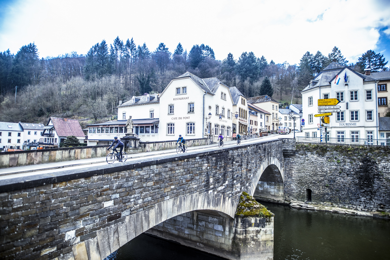 Een groep wielrenners komt fietsend over een brug, het dorp uit, omringd door groen en water.